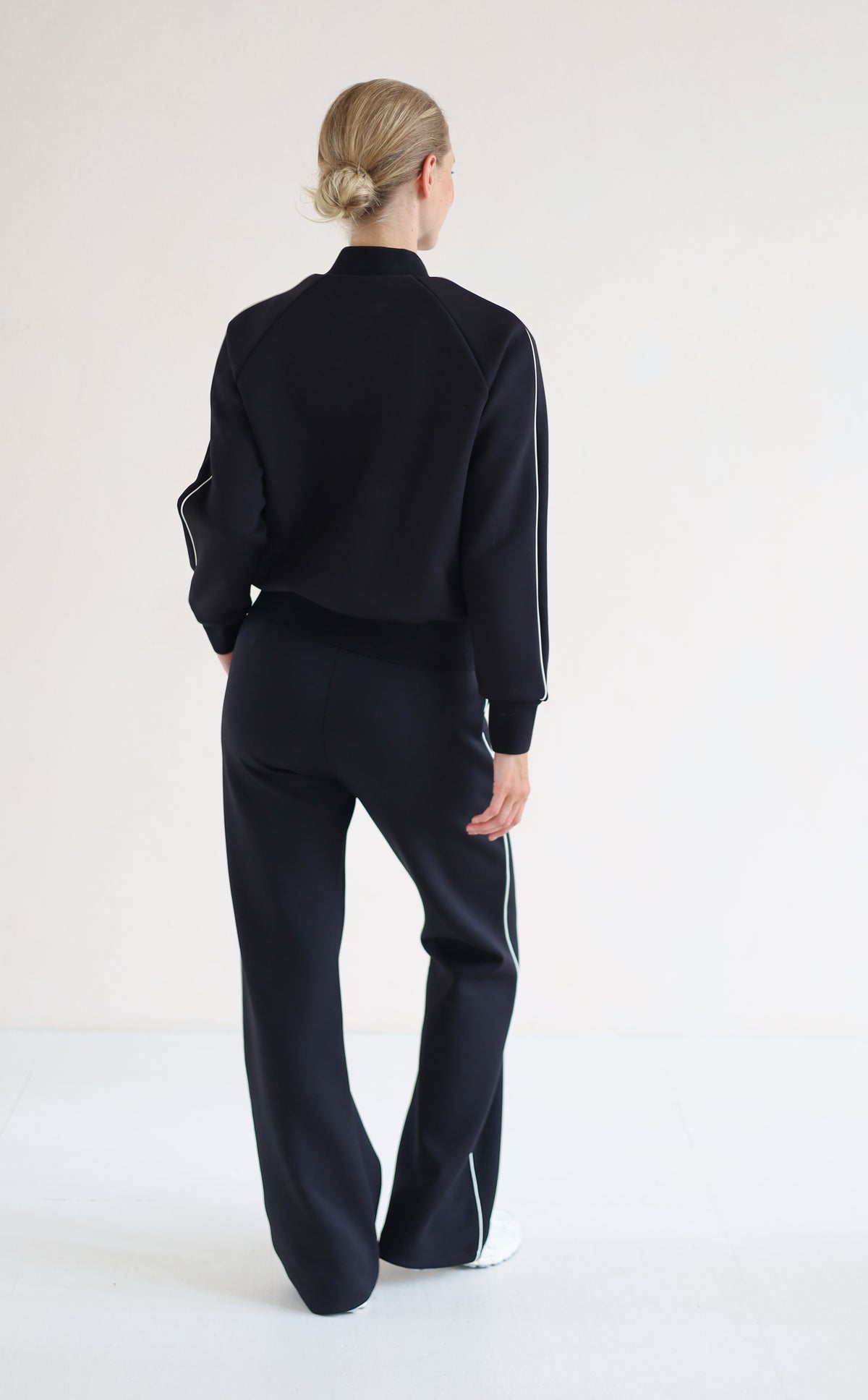 A woman viewed from behind wearing a blackbomber jacket with white side stripes and matching black wide-leg trousers with white side stripes, standing against a plain white studio background.