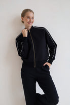 A woman wearing a black zip-up bomber jacket with white piping and a gold zipper, paired with black wide leg trouser, posing against a plain off-white wall.