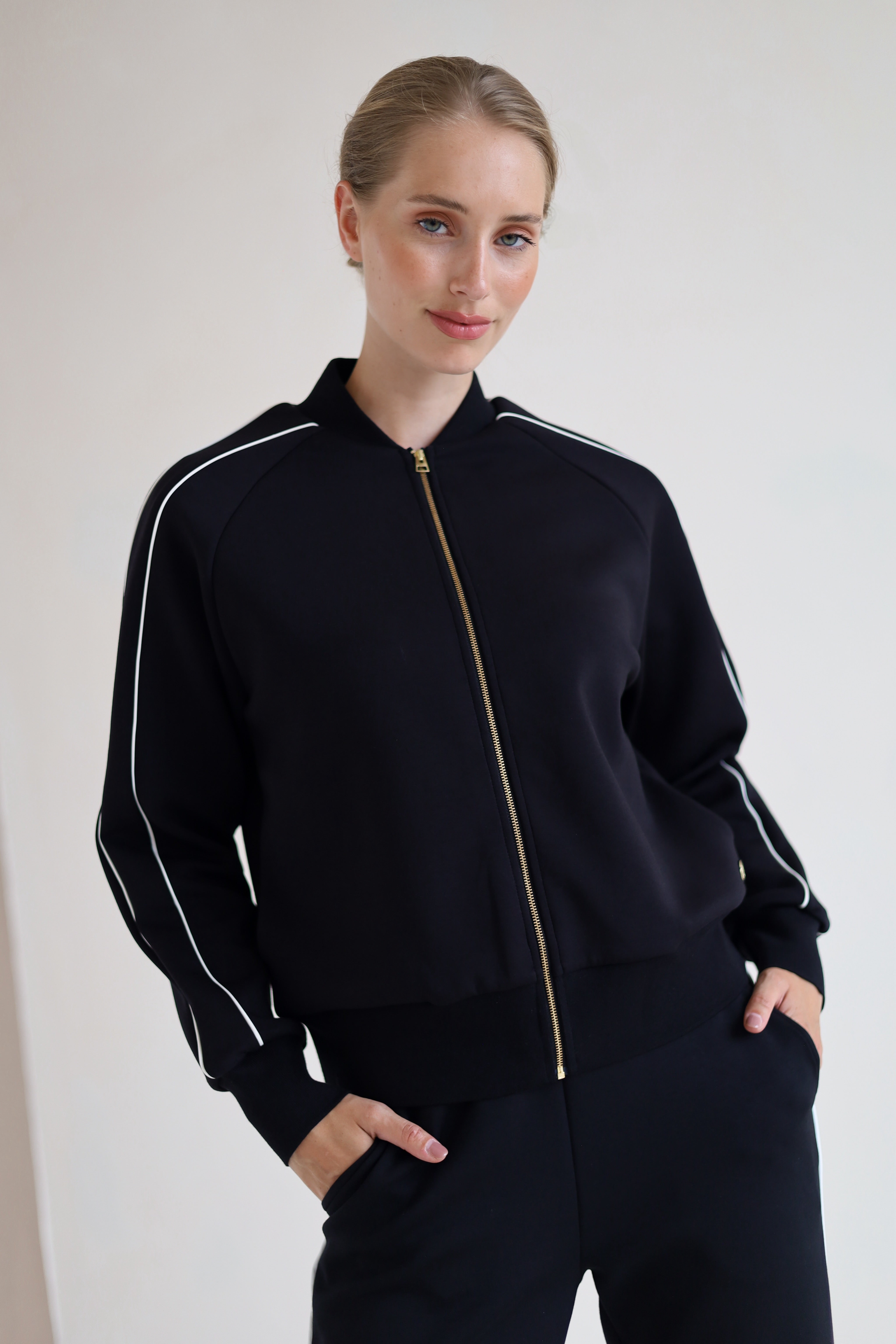 A woman wearing a black zip-up bomber jacket with white piping and a gold zipper, paired with black wide leg trouser, posing against a plain off-white wall.