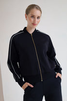 A woman wearing a black zip-up bomber jacket with white piping and a gold zipper, paired with black wide leg trouser, posing against a plain off-white wall.