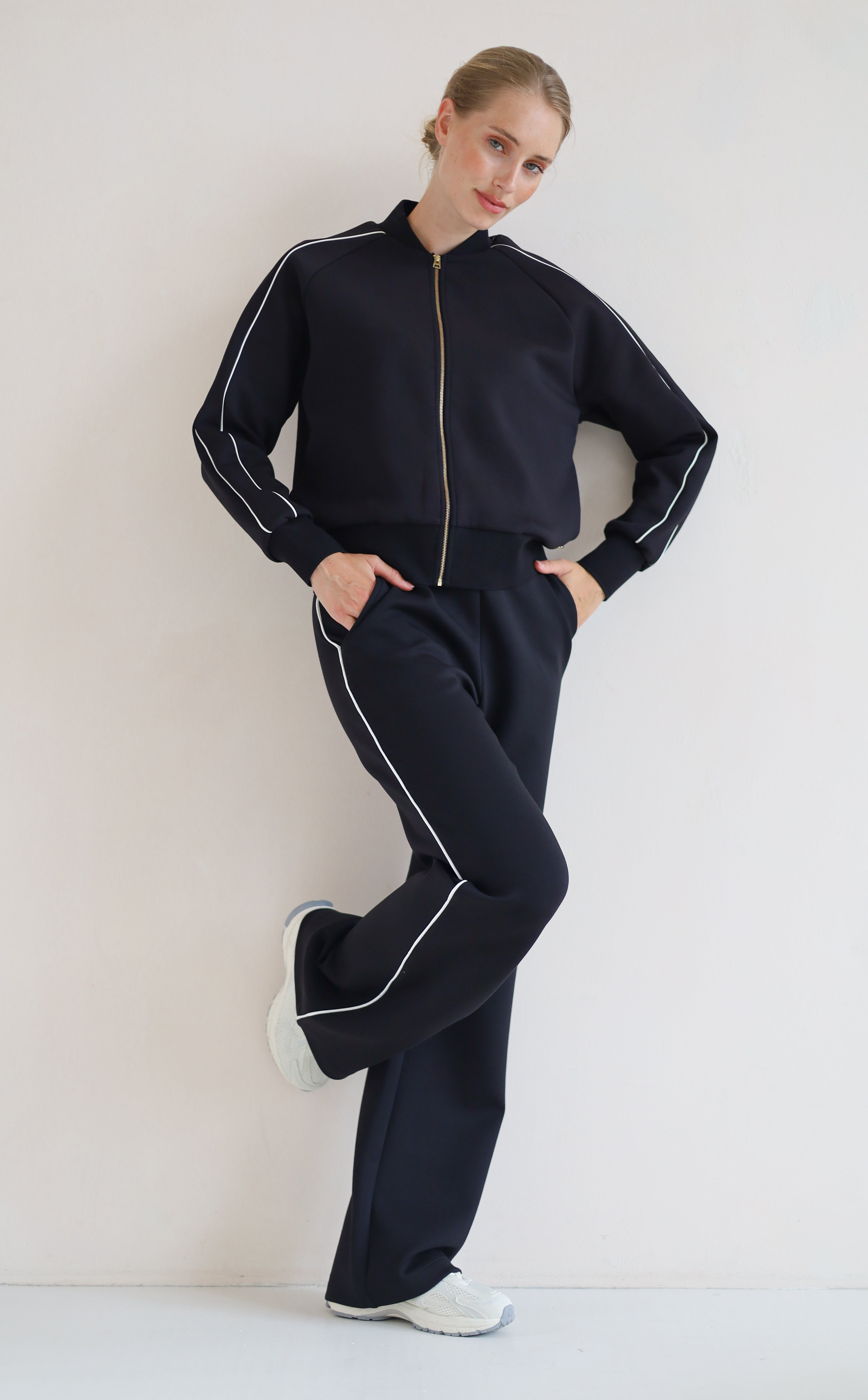A woman modeling a matching black set with white piping details, featuring a zip-up bomber jacket and wide-leg pants, paired with white sneakers against a plain off-white background.