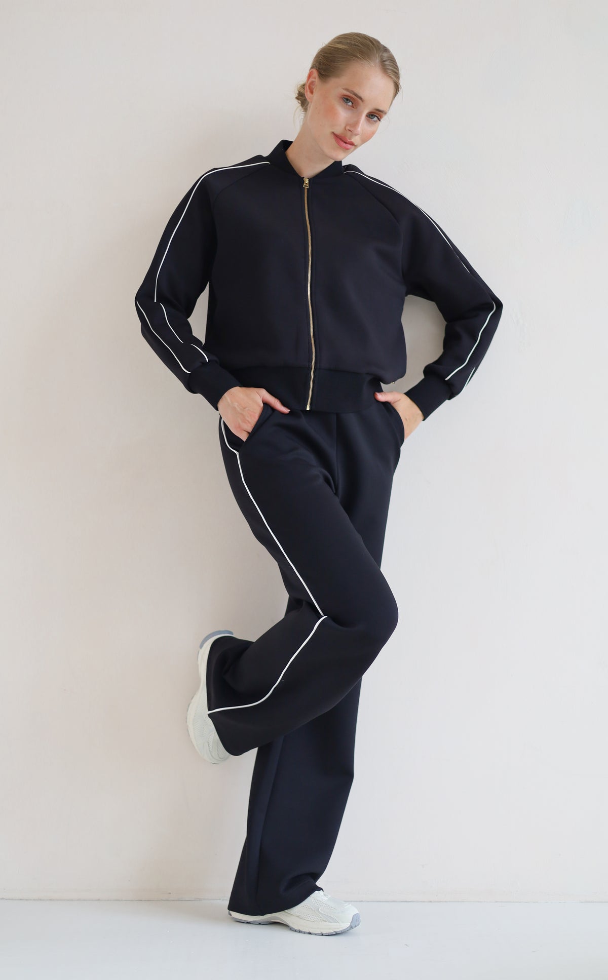 A woman modeling a matching black set with white piping details, featuring a zip-up bomber jacket and wide-leg pants, paired with white sneakers against a plain off-white background.