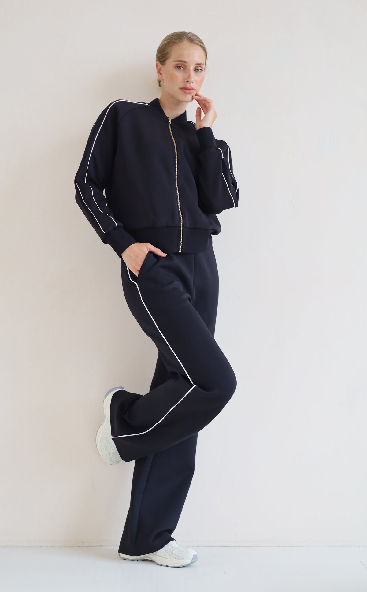 A woman modeling a matching black set with white piping details, featuring a zip-up bomber jacket and wide-leg pants, paired with white sneakers against a plain off-white background.