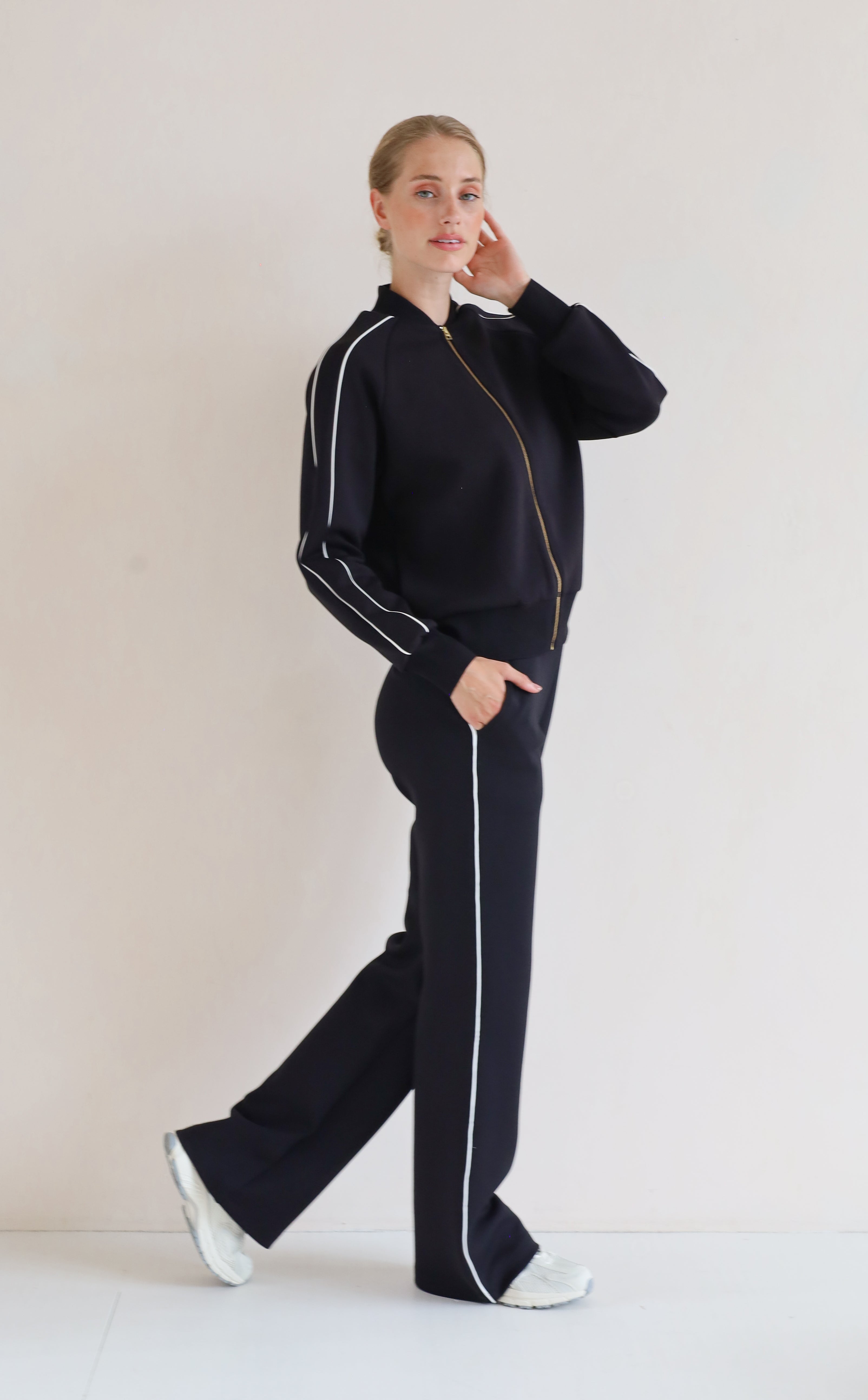 A woman modeling a matching black set with white piping details, featuring a bomber jacket and wide-leg pants, set against a plain off-white studio background.