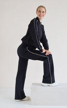 A woman modeling a matching black set with white piping details, posing on a white block against a plain white studio background.