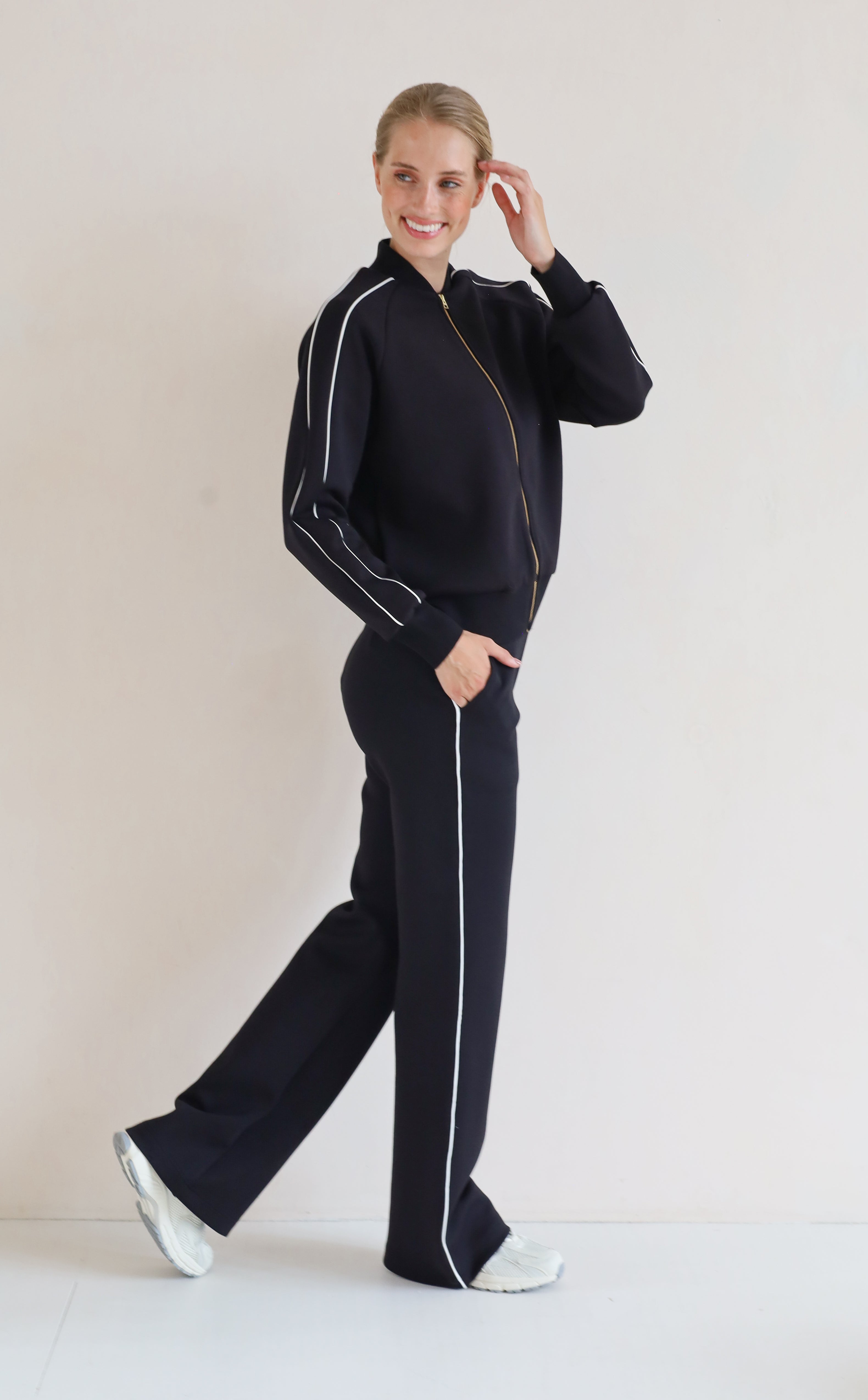 A woman modeling a matching black se with white piping details, featuring a bomber jacket and wide-leg pants, set against a plain light background.