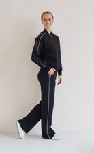 A woman modeling a matching black set with white piping details, consisting of a bomber jacket and wide-leg pants, standing against a plain off-white background.