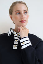A woman wearing a black knit sweater with a contrasting black and white striped collar and striped cuffs, accented with gold buttons, set against a plain light background.