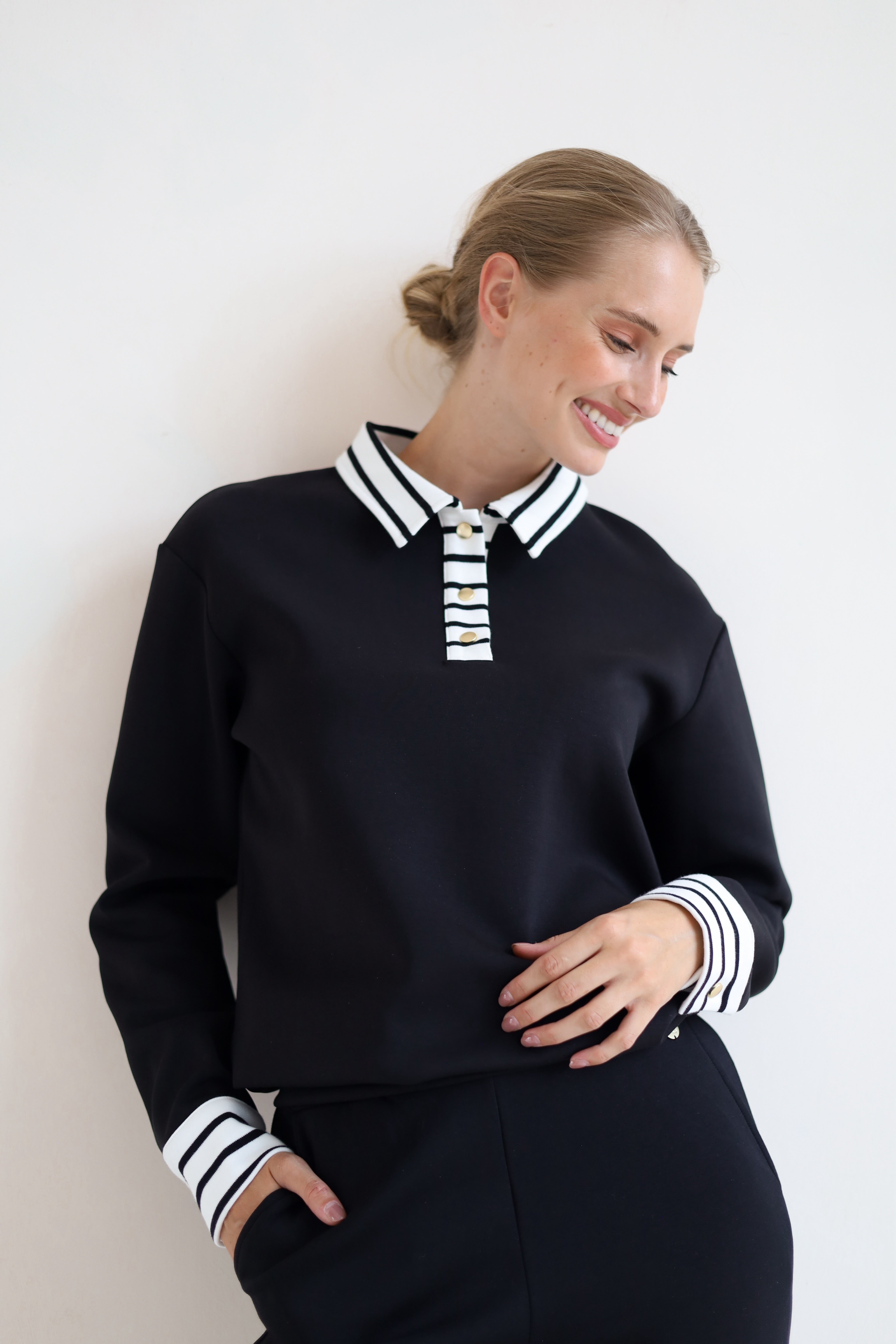 A woman wearing a black long-sleeve knit top with a white and black striped polo-style collar and striped cuffs, paired with black bottoms, posing against a plain white wall.