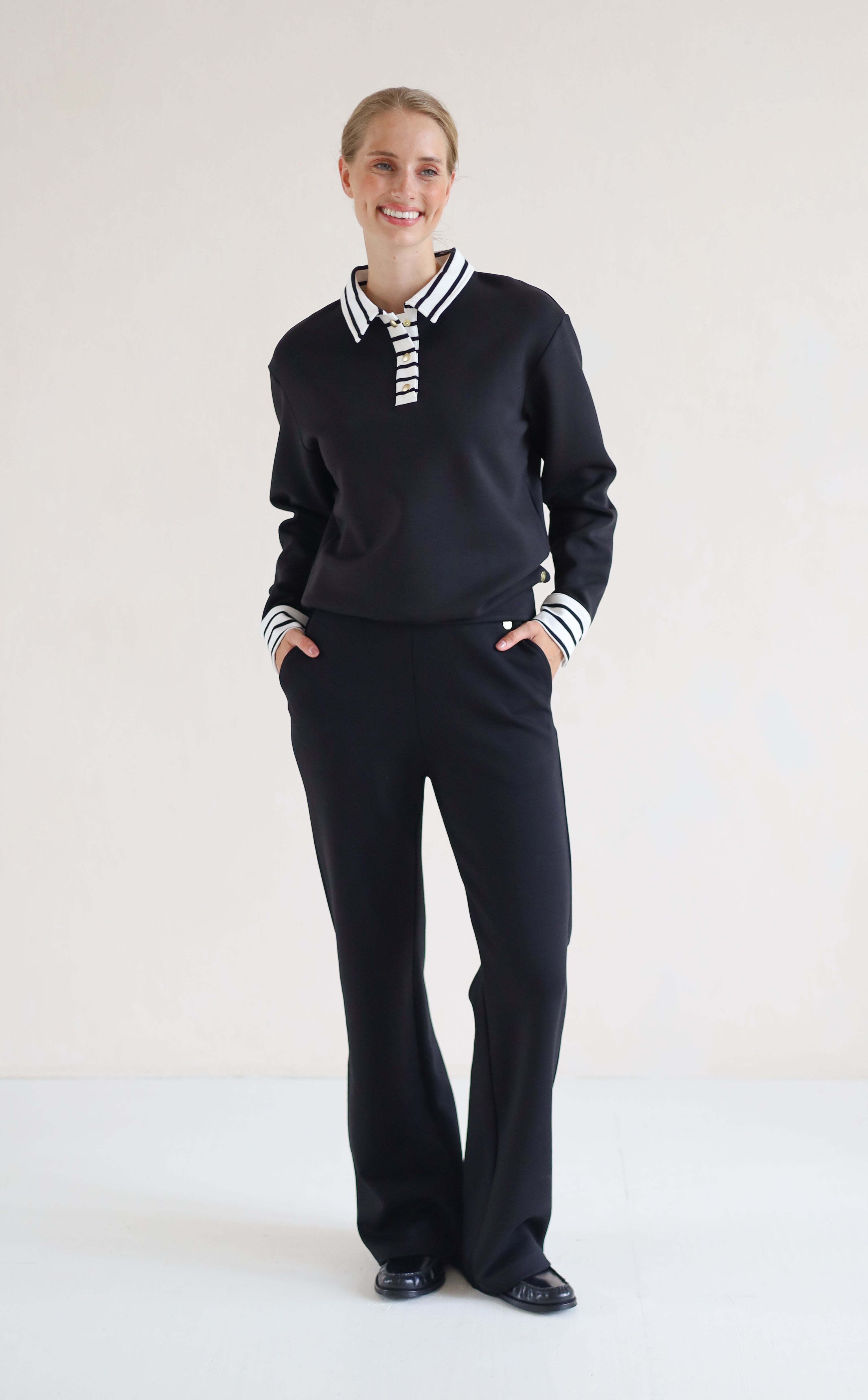 A woman modeling a black long-sleeve polo-style top with a white striped collar and striped cuffs, paired with black wide-leg trousers, against a plain white studio background.