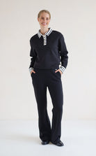 A woman modeling a black long-sleeve polo-style top with a white striped collar and striped cuffs, paired with black wide-leg trousers, against a plain white studio background.