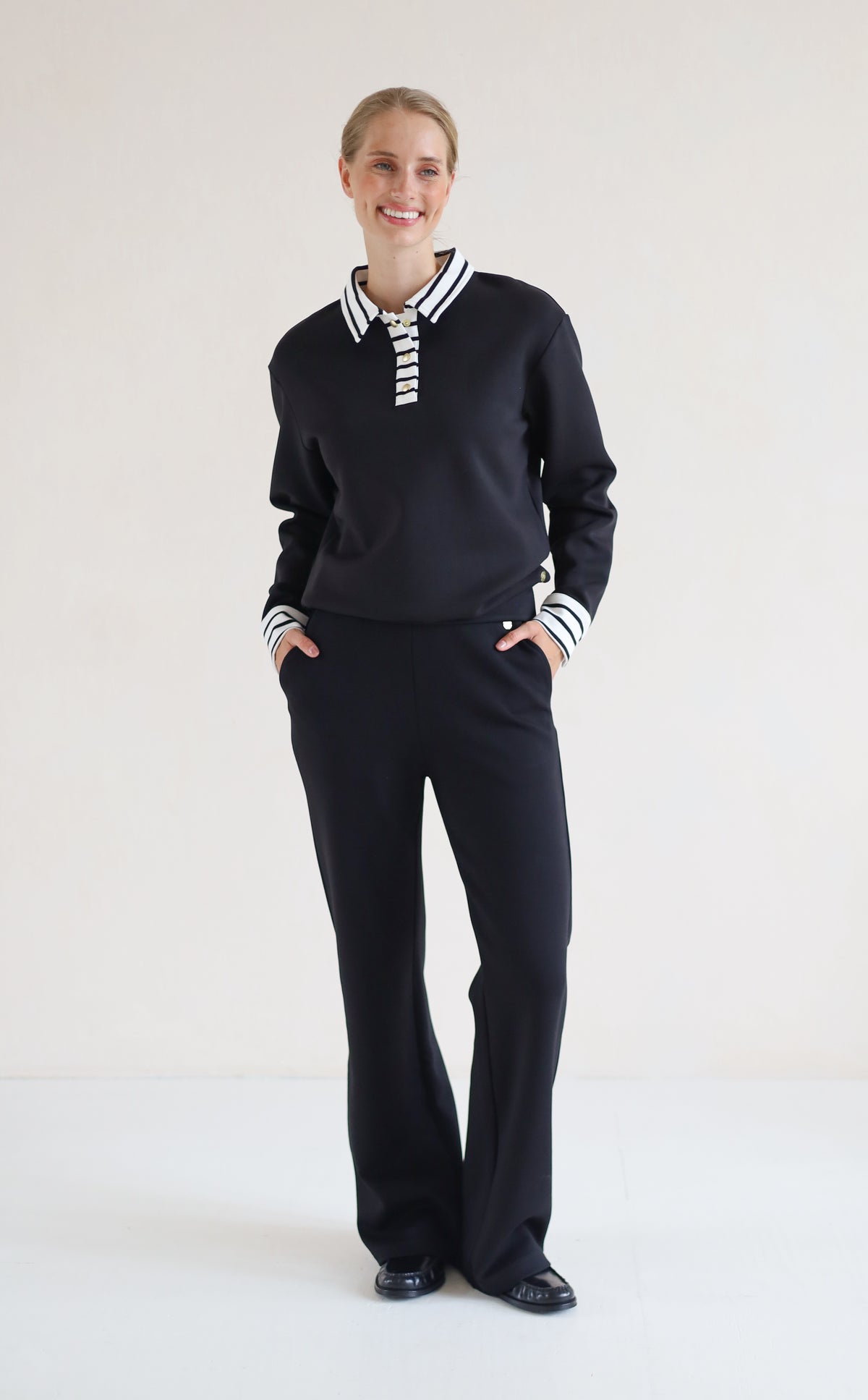 A woman modeling a black long-sleeve polo-style top with a white striped collar and striped cuffs, paired with black wide-leg trousers, against a plain white studio background.