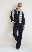 A woman modeling a black knit set consisting of a long-sleeve collared top with black and white horizontal stripes on the shoulders and cuffs, matching black wide-leg trousers, and black shoes, set against a plain off-white wall.