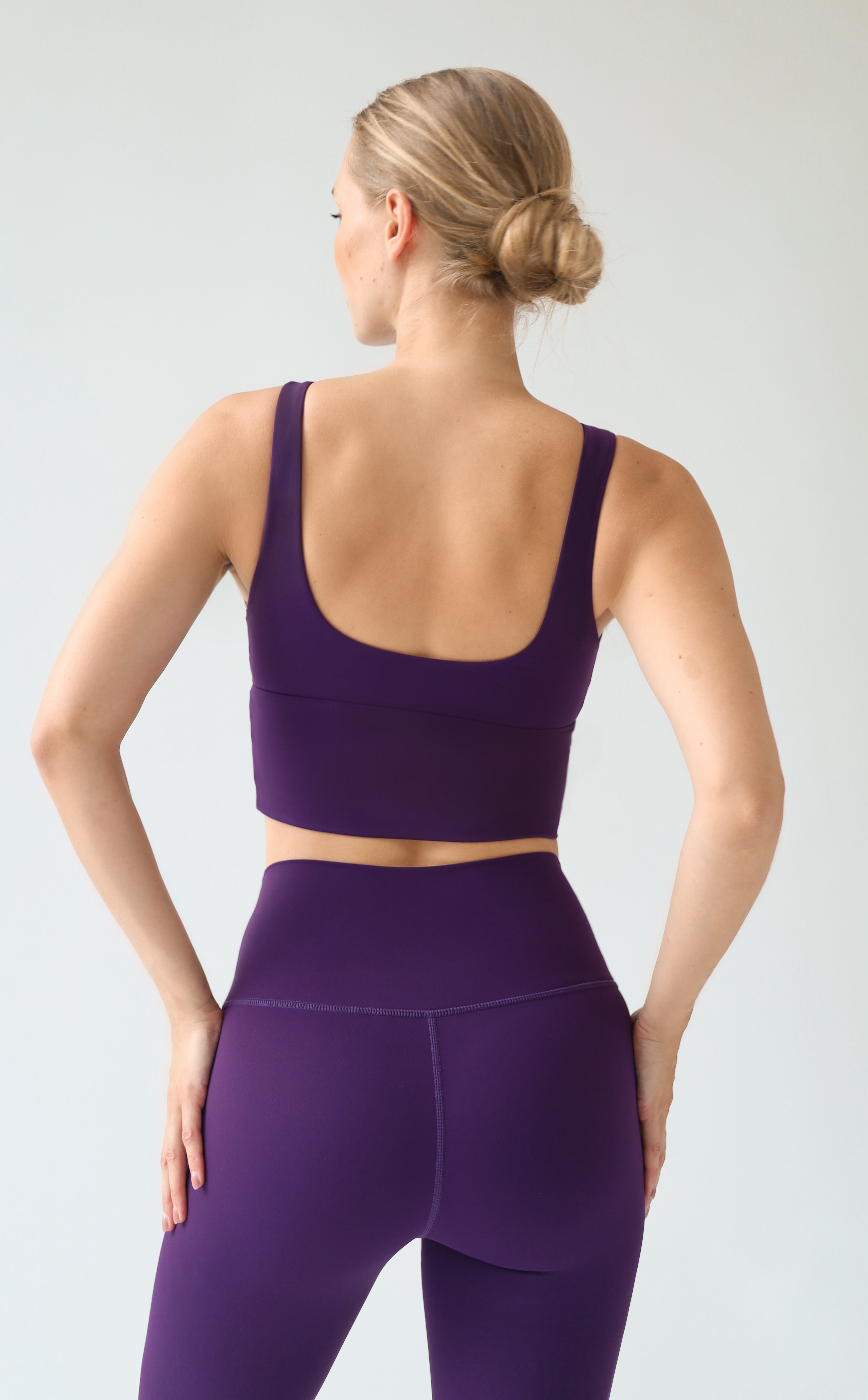A woman shown from the back wearing a matching purple sports bra and high-waisted leggings set against a light gray studio background.