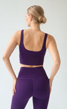 A woman shown from the back wearing a matching purple sports bra and high-waisted leggings set against a light gray studio background.
