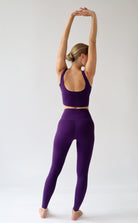 A woman wearing a matching purple activewear set consisting of a sports bra and high-waisted leggings, standing with her arms raised overhead against a light gray studio background.