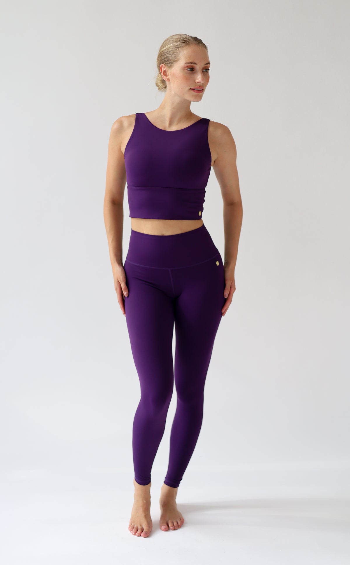 A woman modeling a matching purple activewear set consisting of a sleeveless crop top and high-waisted leggings, standing against a plain light gray studio background.