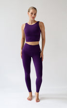 A woman modeling a matching purple activewear set consisting of a sleeveless crop top and high-waisted leggings, standing against a plain light gray studio background.