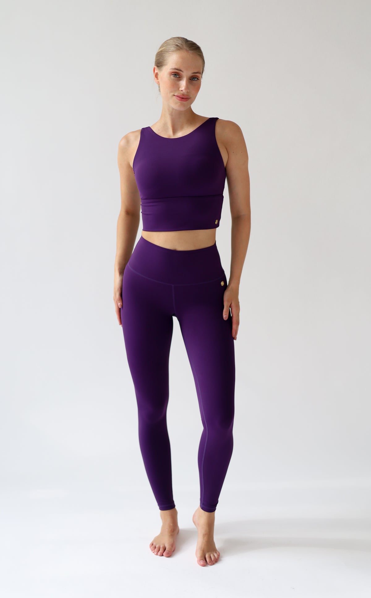 A woman modeling a matching purple activewear set consisting of a sleeveless crop top and high-waisted leggings, standing against a plain light gray studio background.