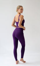 A woman modeling a matching purple activewear set consisting of a sports bra and high-waisted leggings, shown from a side-back angle against a plain white studio background.