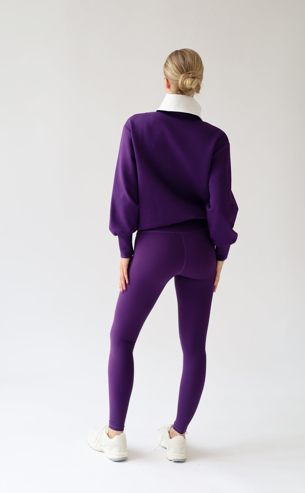 A woman viewed from behind wearing a matching purple cardigan and leggings, with a light grey collar detail on the cardigan and white sneakers, against a plain light gray studio background.