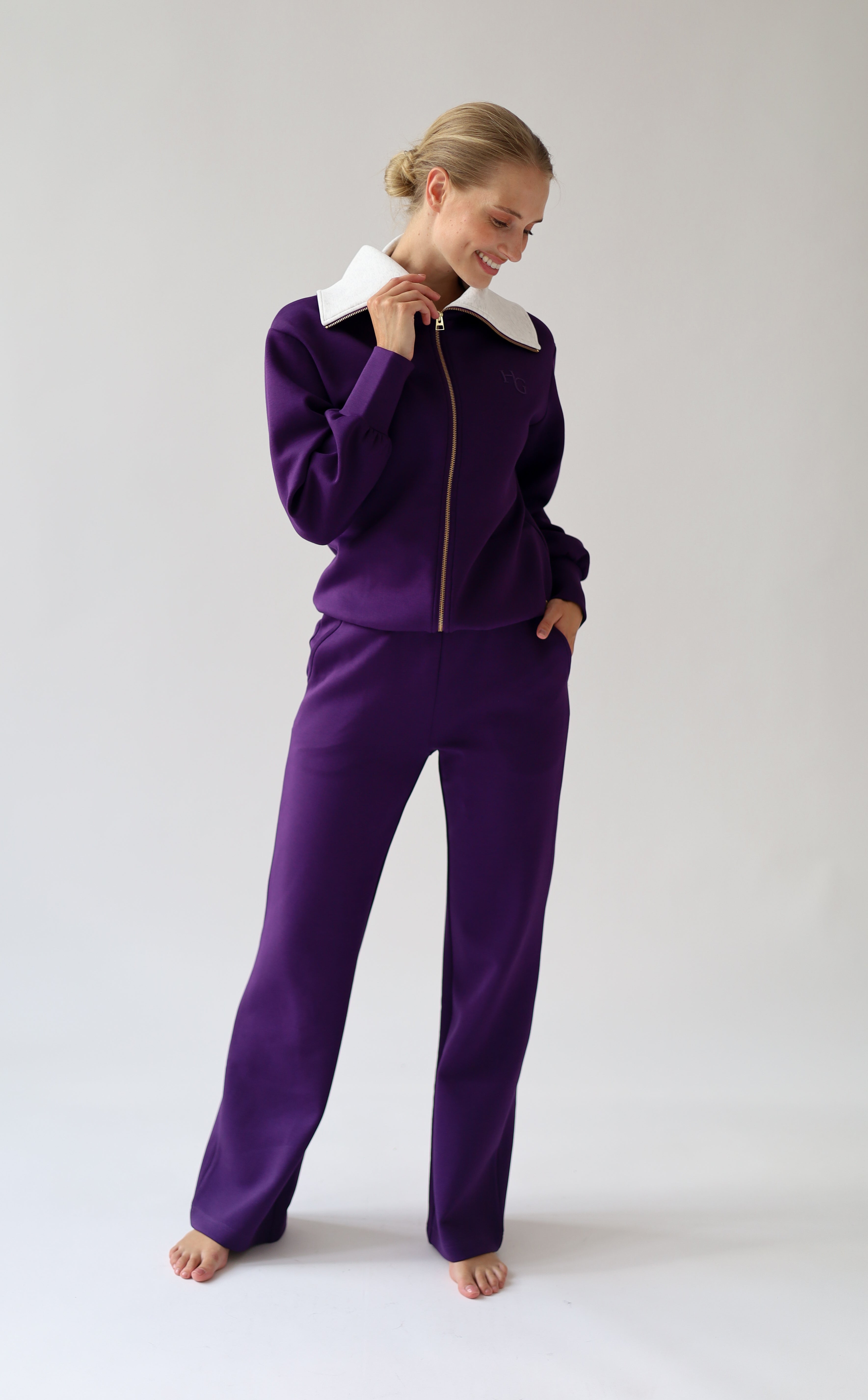 A woman modeling a purple city wear with a light grey collar and gold zipper, standing against a light gray studio background.
