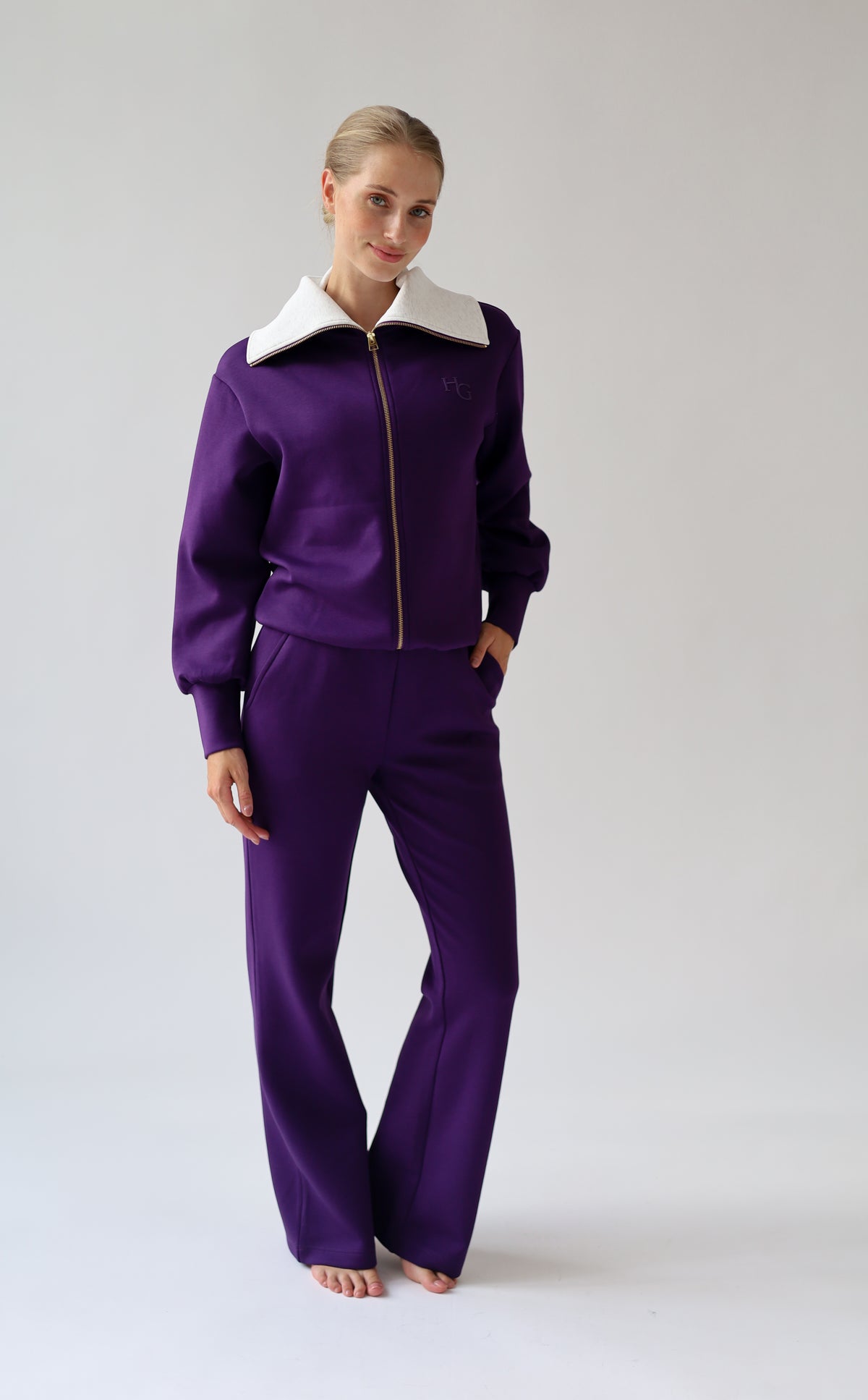 A woman modeling a purple city wear with a light grey collar and gold zipper, standing against a light gray studio background.