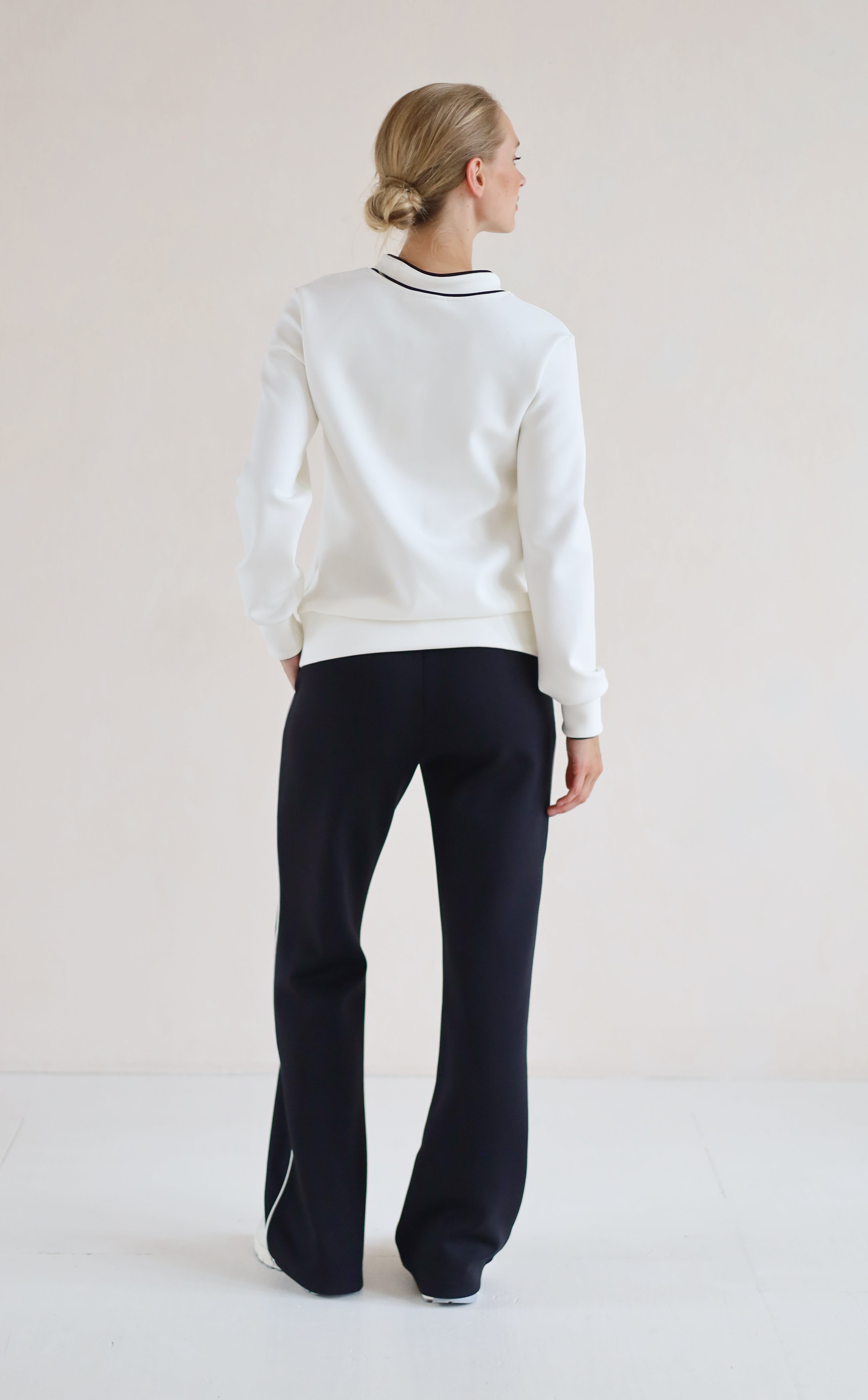 A woman is shown from the back wearing a white v-neck sweater with black trim and black wide-leg trousers, standing against a plain light beige studio background.