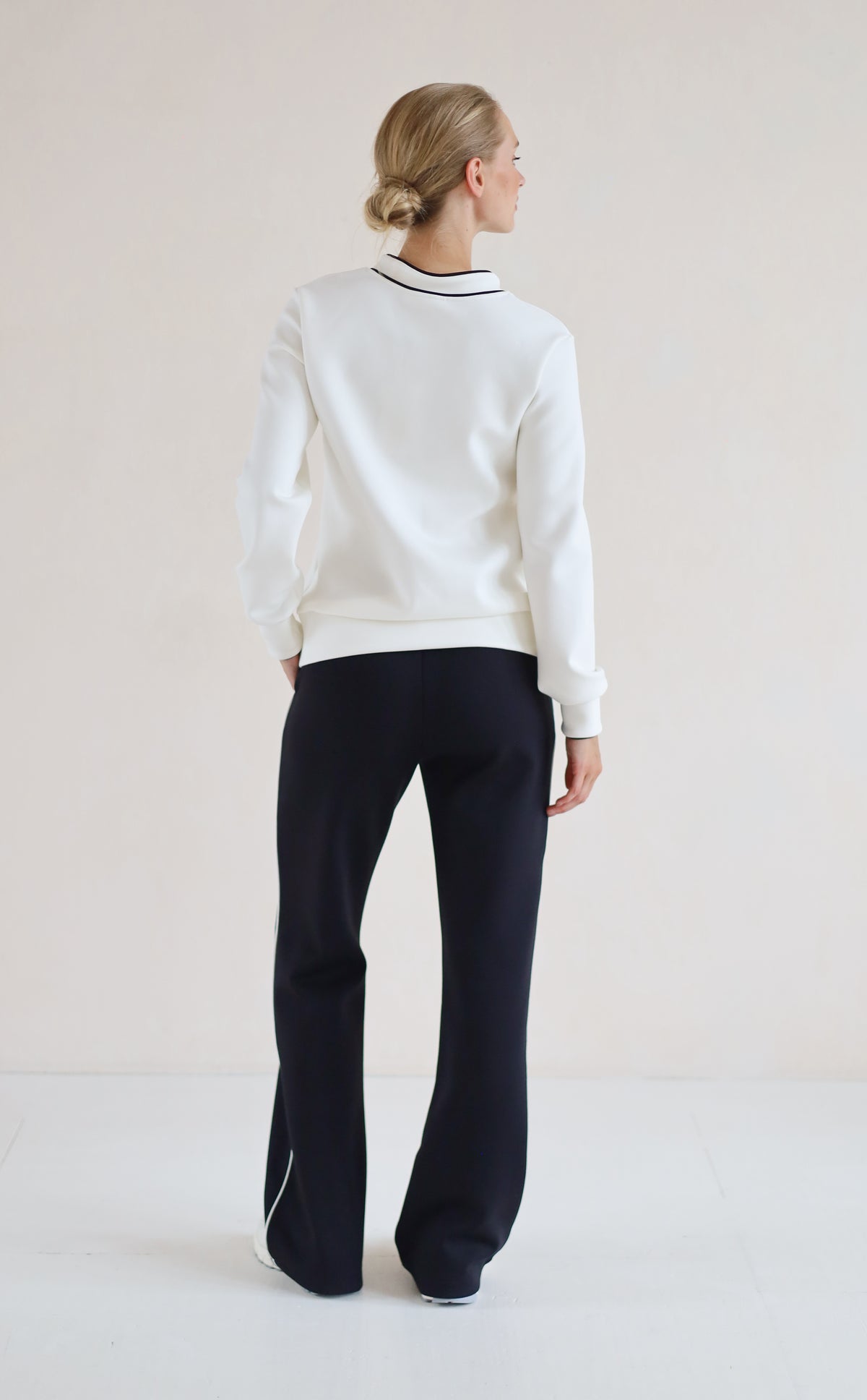 A woman is shown from the back wearing a white v-neck sweater with black trim and black wide-leg trousers, standing against a plain light beige studio background.