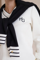 House of Gravity V-Neck Sweater in Off-White Marble and Black, close-up of the embroidered HG logo, styled with the Button Polo Sweater in Black draped over the shoulders.