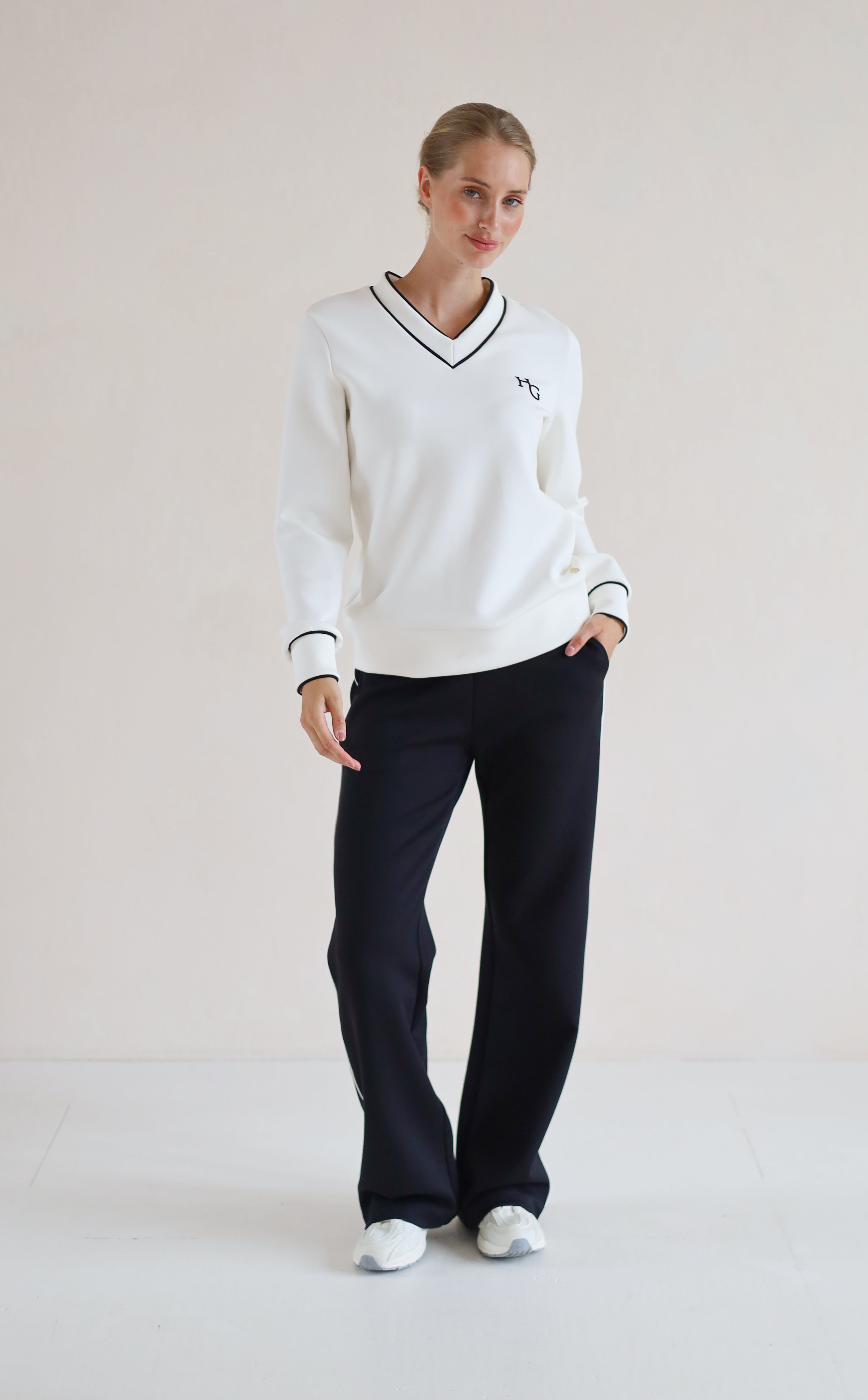 A woman modeling a white long-sleeve V-neck sweater with black trim and a small embroidered chest logo, paired with black wide-leg pants and white sneakers, against a plain light beige studio background.