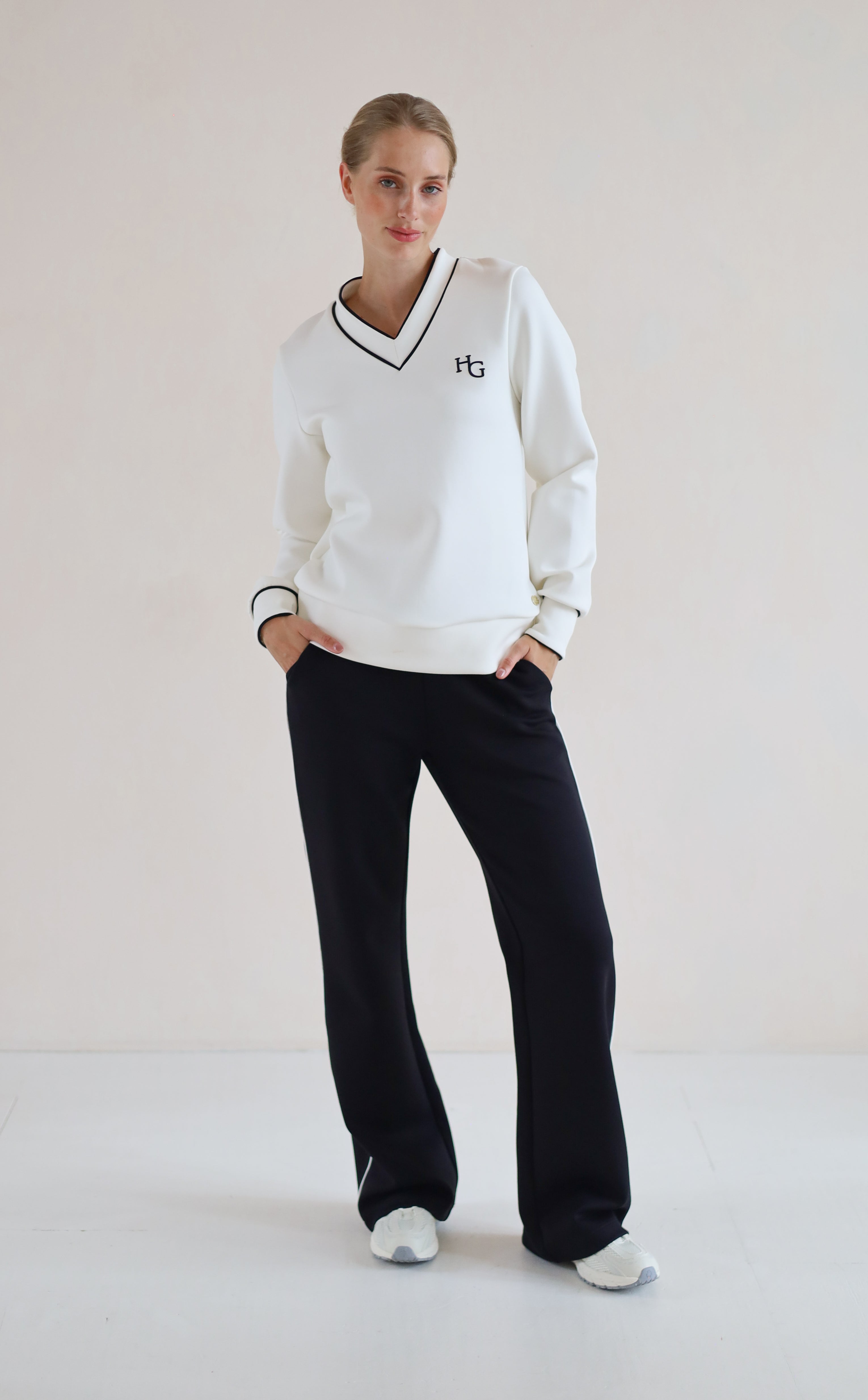 A woman modeling a white long-sleeve V-neck sweatshirt with black trim and a small black 'hg' logo on the chest, paired with black wide-leg sweatpants and white sneakers, set against a plain off-white studio background.