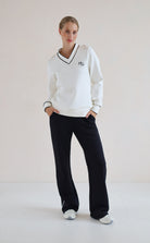 A woman modeling a white long-sleeve V-neck sweatshirt with black trim and a small black 'hg' logo on the chest, paired with black wide-leg sweatpants and white sneakers, set against a plain off-white studio background.