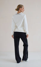 A woman shown from the back wearing a white long-sleeve sweatshirt with a black and white striped collar, paired with black wide-leg trouser featuring thin white side stripes, set against a plain light beige studio background.