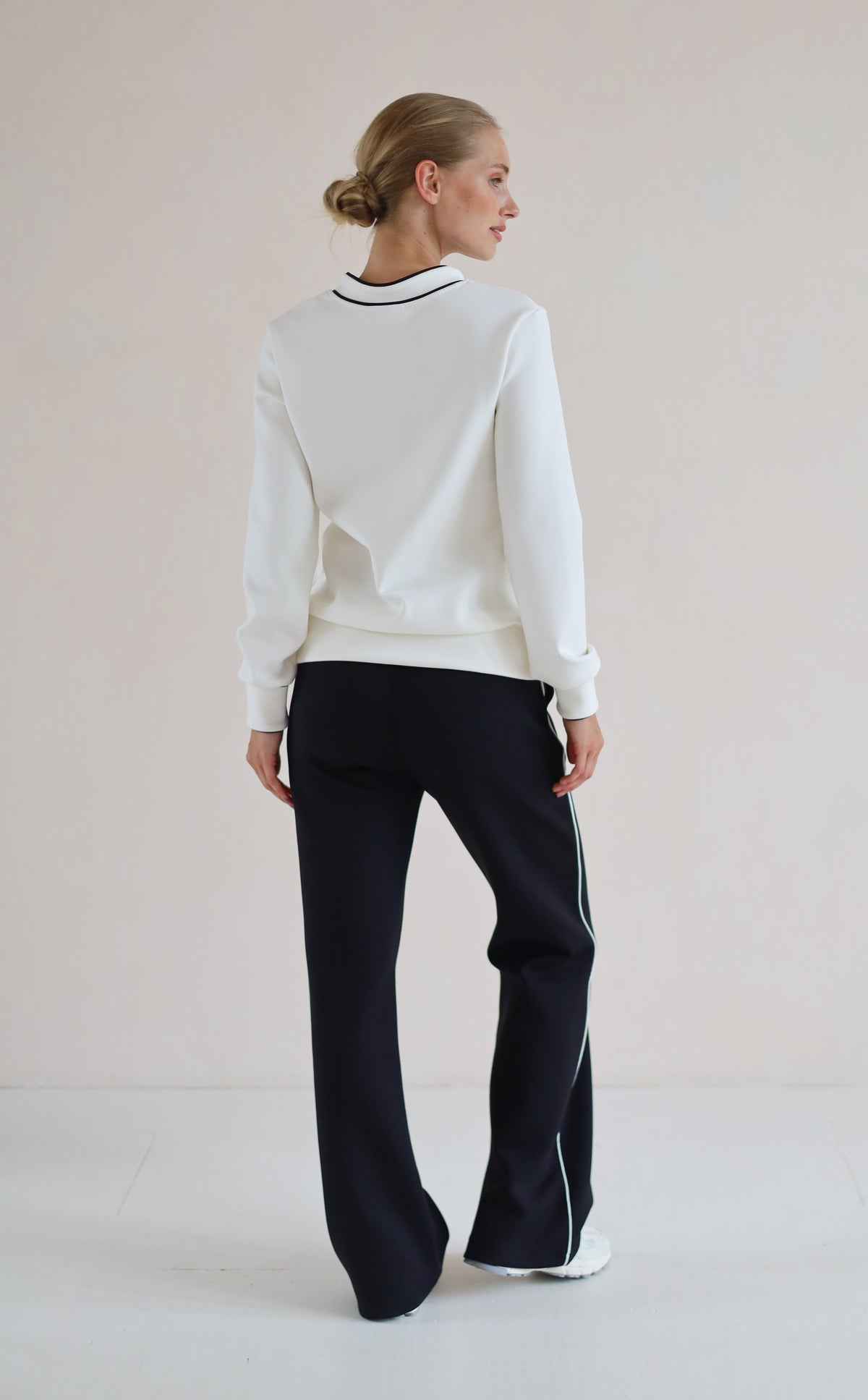 A woman shown from the back wearing a white long-sleeve sweatshirt with a black and white striped collar, paired with black wide-leg trouser featuring thin white side stripes, set against a plain light beige studio background.