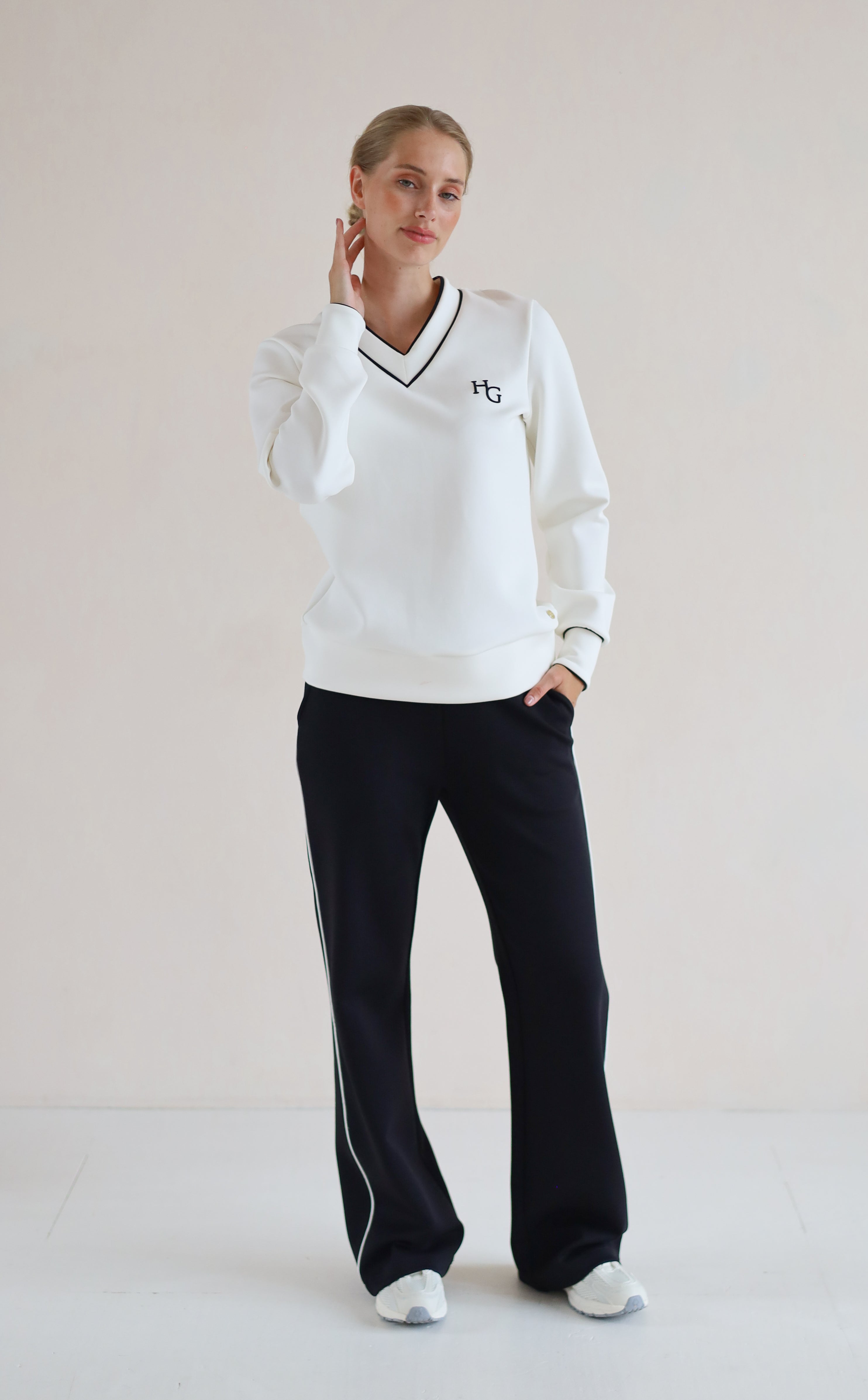 A woman modeling a white long-sleeve V-neck sweatshirt with black trim and a small black monogram logo on the left chest, paired with black wide-leg trouser featuring white side stripes, against a plain light beige studio background.