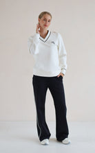 A woman modeling a white long-sleeve V-neck sweatshirt with black trim and a small black monogram logo on the left chest, paired with black wide-leg trouser featuring white side stripes, against a plain light beige studio background.