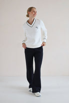 A woman modeling a white long-sleeve V-neck sweater with black trim and a small embroidered logo on the chest, paired with black pants and white sneakers, in a minimalist studio setting.