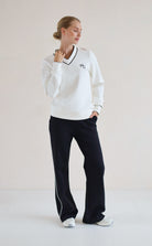 A woman modeling a white long-sleeve V-neck sweatshirt with black trim and a small embroidered logo on the chest, paired with black wide-leg trouser and white sneakers, against a plain light beige studio background.