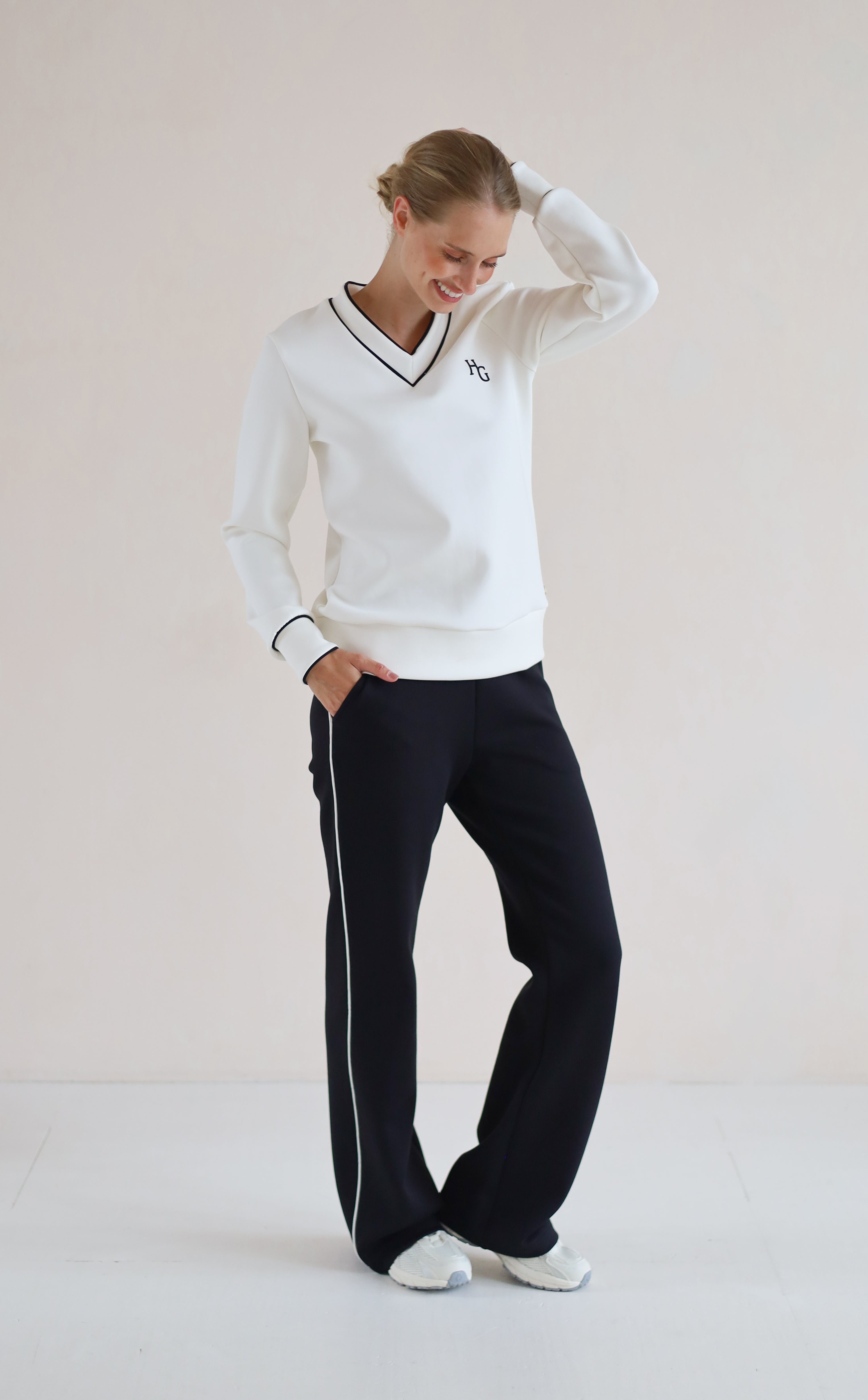 A woman modeling a white long-sleeve V-neck sweatshirt with black trim and a small embroidered logo on the chest, paired with black trouser featuring a white side stripe, against a plain light beige studio background.