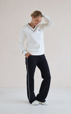 A woman modeling a white long-sleeve V-neck sweatshirt with black trim and a small embroidered logo on the chest, paired with black trouser featuring a white side stripe, against a plain light beige studio background.