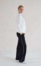 A woman stands in profile wearing a white long-sleeve V-Neck Sweater and black wide-leg pants with a white side stripe, against a plain light beige studio background.