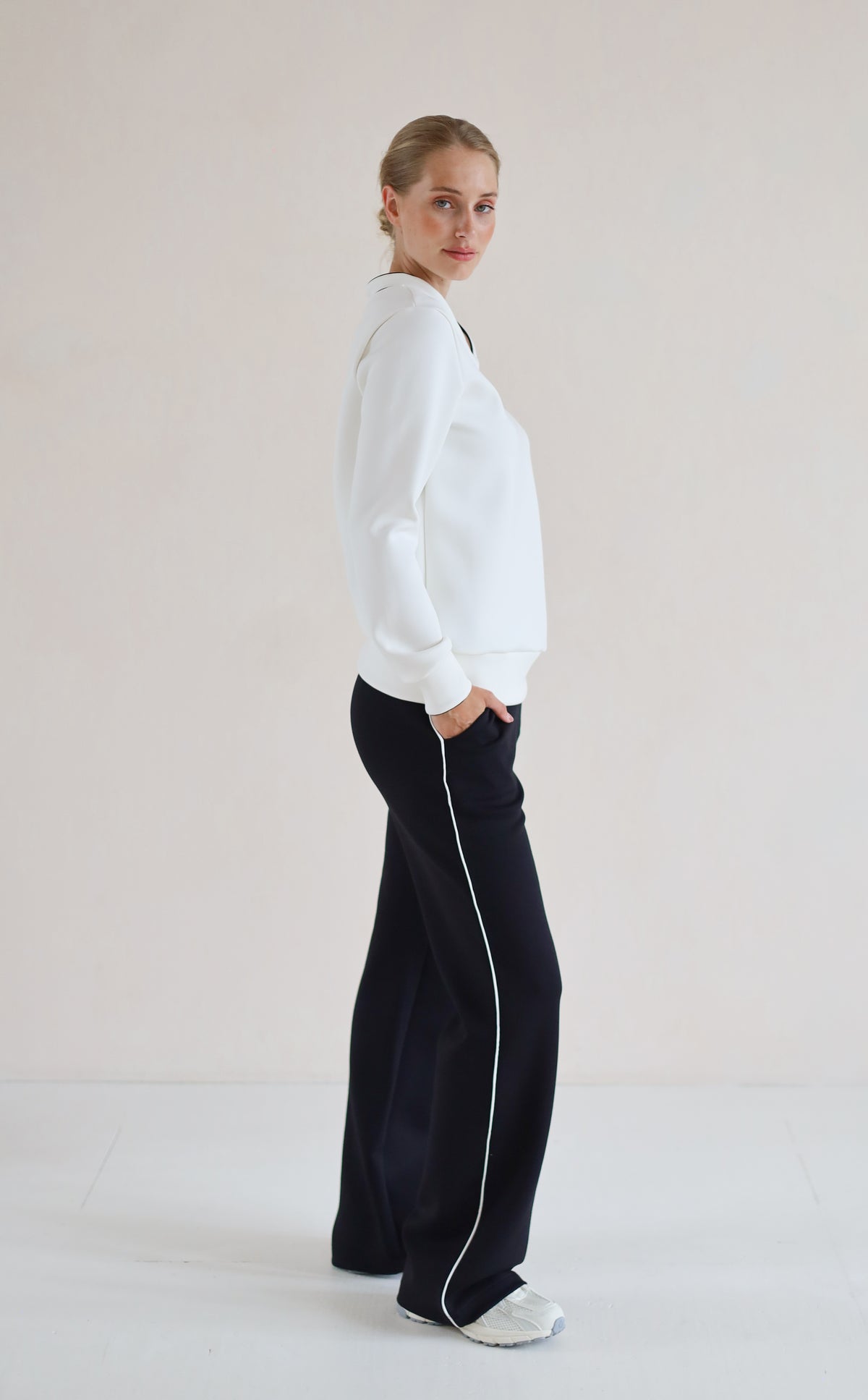 A woman stands in profile wearing a white long-sleeve V-Neck Sweater and black wide-leg pants with a white side stripe, against a plain light beige studio background.