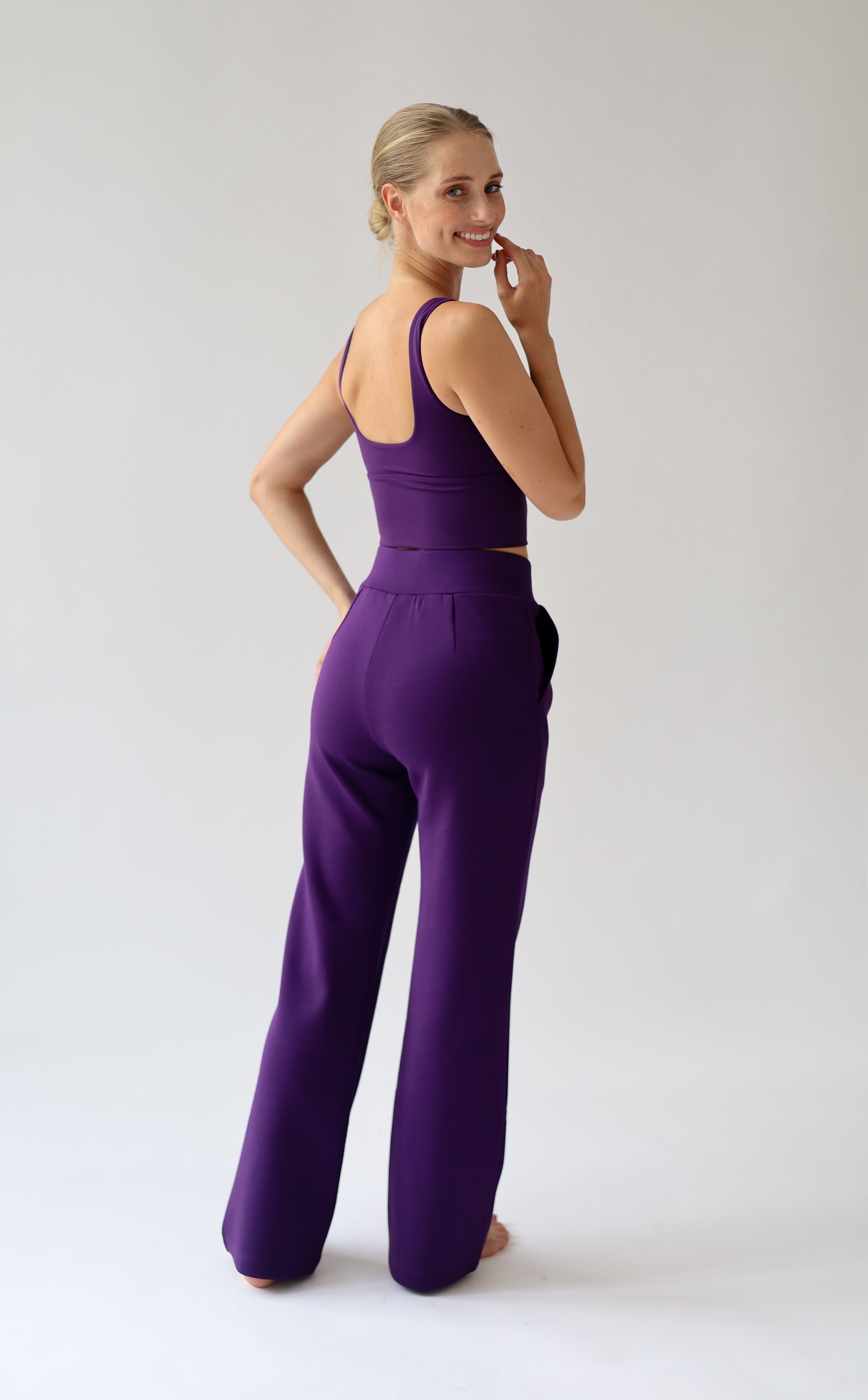 A woman modeling a matching purple set set consisting of a sleeveless crop top and high-waisted wide-leg pants, shown from a three-quarters back view against a light gray studio background.