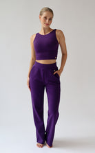 A woman modeling a matching purple activewear set consisting of a sleeveless crop top and high-waisted wide leg trousers, standing against a light gray studio background.