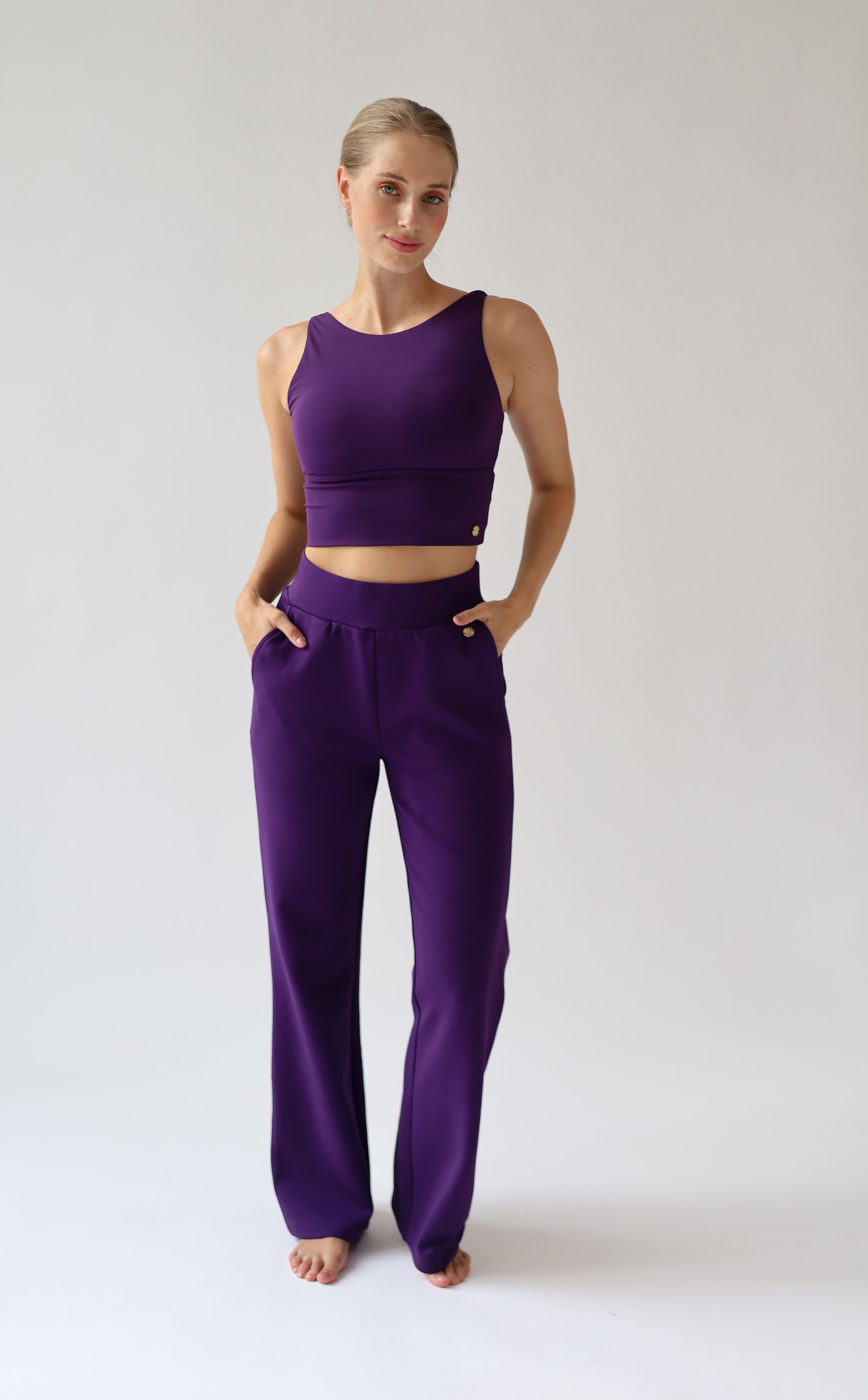 A woman modeling a matching purple activewear set consisting of a sleeveless crop top and high-waisted wide leg trousers, standing against a light gray studio background.